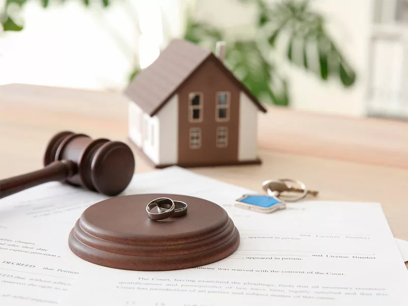 Gavel laying on a table next to a model home and wedding rings